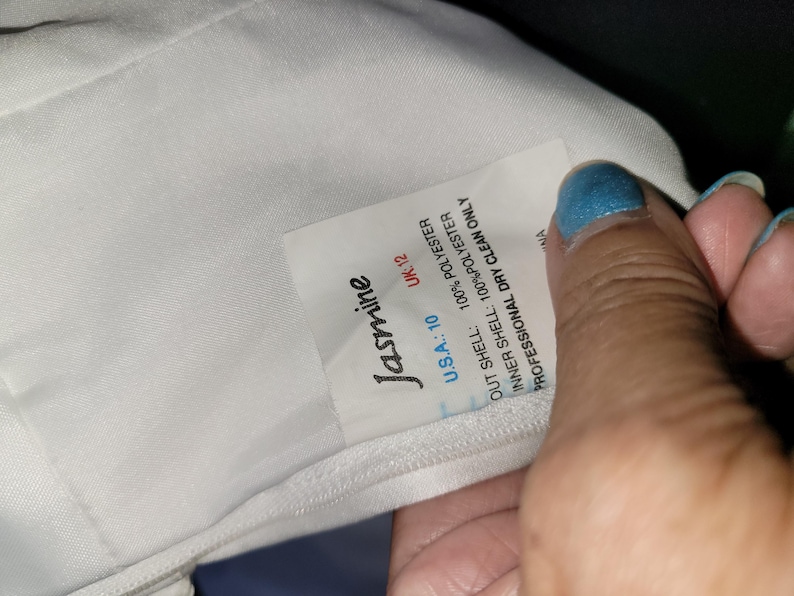 Może przedstawiać: Wnętrze białej odzieży z wszytą metką z nazwą marki Jasmine. Metka zawiera informacje: U.S.A.: 10, UK: 12, 100% poliester dla warstwy zewnętrznej i wewnętrznej oraz instrukcję dotyczącą wyłącznie profesjonalnego czyszczenia chemicznego.