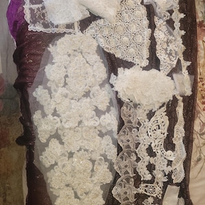 May include: An assortment of white lace fabric pieces, including floral appliques, trim, and decorative panels. The lace varies in pattern and texture, with some pieces featuring beaded embellishments. The fabrics are arranged on a dark, patterned surface.