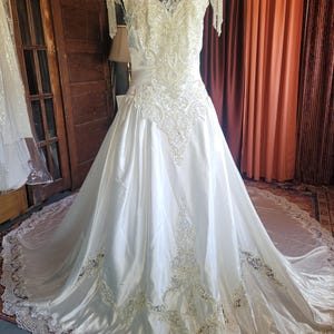 May include: A white wedding dress with a high neckline and a full skirt. The bodice is embellished with intricate lace and beadwork. The skirt flows into a long train with lace trim. The dress is displayed on a mannequin.