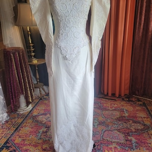 May include: Ivory-colored wedding dress with a fitted bodice and a flowing skirt. The bodice features intricate lace detailing and a V-neckline. The long sleeves have a unique, puffed design. The dress is displayed on a black mannequin.