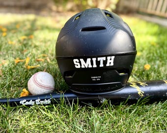 May include: A black baseball helmet with the name "SMITH" in white lettering, a baseball, and a black baseball bat with the name "Cody Smith" in white lettering, all resting on green grass.