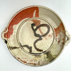 May include: A round ceramic serving platter with two side handles, featuring a cream-coloured base with abstract terracotta orange and dark brown brushstroke patterns. The surface has a textured, glazed finish with visible variations in colour.