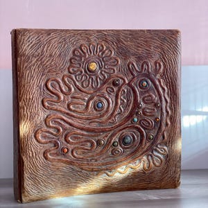 May include: A brown, square book with a textured cover. The cover features a raised, ornate design of a bird-like figure and sun, with colourful stone accents. The book is displayed on a light grey surface.