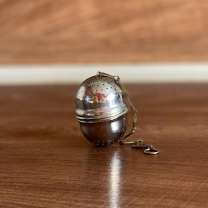 May include: Silver tea infuser with a chain and a round, perforated design. The infuser is on a wooden surface.