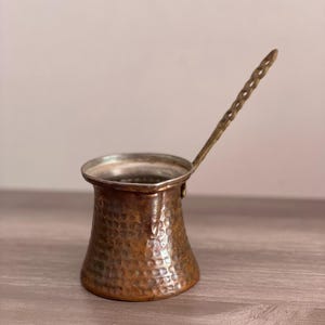 May include: A hammered copper Turkish coffee pot with a brass handle. The pot has a traditional design and is perfect for brewing strong coffee.