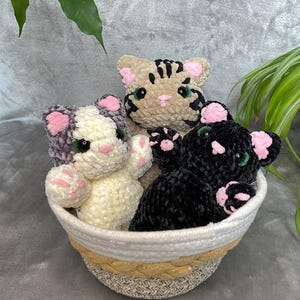Peut inclure: Trois jouets chats en crochet peluche dans un panier tressé. Un chat est beige avec des rayures noires, un est blanc avec des taches grises, et un est noir uni. Chaque chat a des oreilles roses, un nez rose, des coussinets roses et des yeux verts.