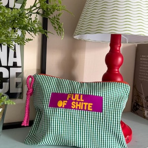 May include: A rectangular zipped pouch featuring a green and white gingham pattern. A bright pink rectangular patch is centred on the front with the text FULL OF SHITE printed in yellow. A pink braided cord is attached to the metal zip pull.