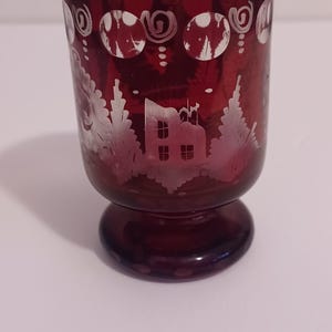 May include: A small, ruby red glass cup with a white etched design of a house and trees. The cup has a round base and a wide rim.