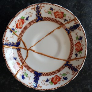 Può includere: Un piatto in ceramica bianca con un motivo floreale e riparazioni kintsugi dorate. Il piatto presenta disegni floreali rossi e blu, un bordo marrone e crepe visibili riempite d'oro. Il piatto ha un bordo smerlato.