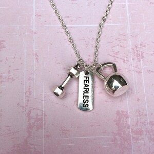 May include: Silver-toned necklace with three charms: a dumbbell, a kettlebell, and a rectangular tag with the word "FEARLESS". The chain is delicate, and the charms are detailed. The background is a light pink, textured surface.