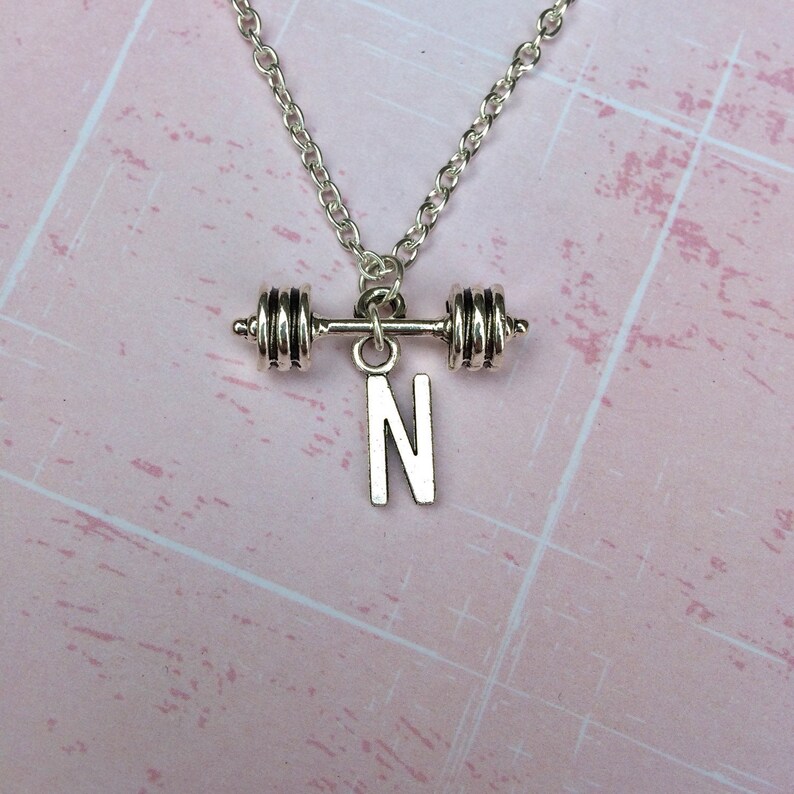 May include: Silver necklace with a barbell charm and a letter N pendant. The necklace is on a pink background.