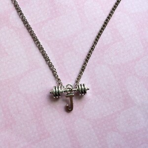 May include: Silver necklace with a barbell charm and the letter J hanging from it.