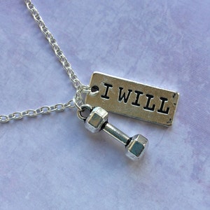 May include: Silver-toned necklace featuring a small dumbbell charm and a rectangular tag with the text "I WILL". The chain is delicate, and the pendant is set against a soft, purple background. A motivational piece for fitness enthusiasts.