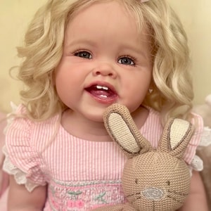 May include: A smiling baby doll with blonde curly hair, wearing a pink striped dress and pink hair bows. The doll is holding a brown knitted bunny toy. The doll has blue eyes and is smiling, showing its teeth.