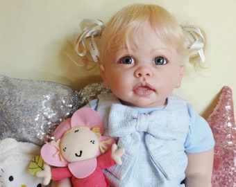 Puede incluir: Una muñeca rubia con ojos azules que lleva un vestido azul claro con un lazo blanco. La muñeca está sentada en una almohada rosa y plateada con lentejuelas.