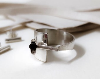May include: A silver ring with a black stone and a geometric design. The ring is on a finger and the background is white.