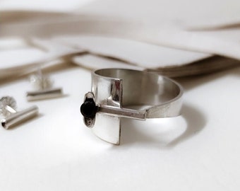 May include: A silver ring with a black stone and a geometric design. The ring is on a finger and the background is white.
