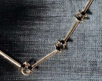 May include: A gold necklace with black accents in the shape of crosses and flowers. The necklace is on a gray textured background.