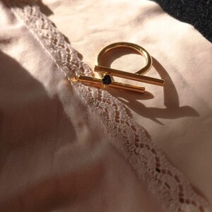 May include: A gold ring with a thick, horizontal bar across the top. The ring is on a light pink fabric with lace trim.
