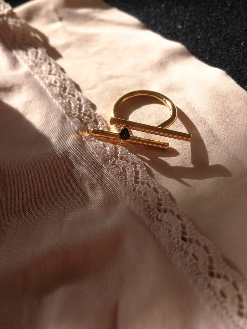 May include: A gold ring with a thick, horizontal bar across the top. The ring is on a light pink fabric with lace trim.