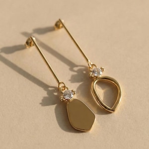 May include: A pair of gold-colored earrings. Each earring features a long, thin post with a small, clear gemstone accent. One earring has a teardrop-shaped pendant, while the other has a solid, geometric shape.