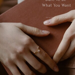 May include: A gold ring with a small, clear stone set in a V-shaped band. The ring is on a person's finger, and the background is a brown book with the text "Dare To Be What You Want" and "Classic Ring" and "Gold Minimalist Ring".