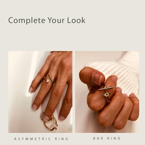 May include: Two gold rings on a hand. The first ring is a simple bar ring with a small clear stone in the center. The second ring is an asymmetric ring with a small clear stone in the center.