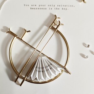 Puede incluir: Un soporte de metal dorado con una parte superior redonda y dos brazos largos. Un abanico de papel blanco se coloca en el soporte. El abanico tiene el texto "You are your only salvation, Awareness is the key." escrito en &eacute;l.