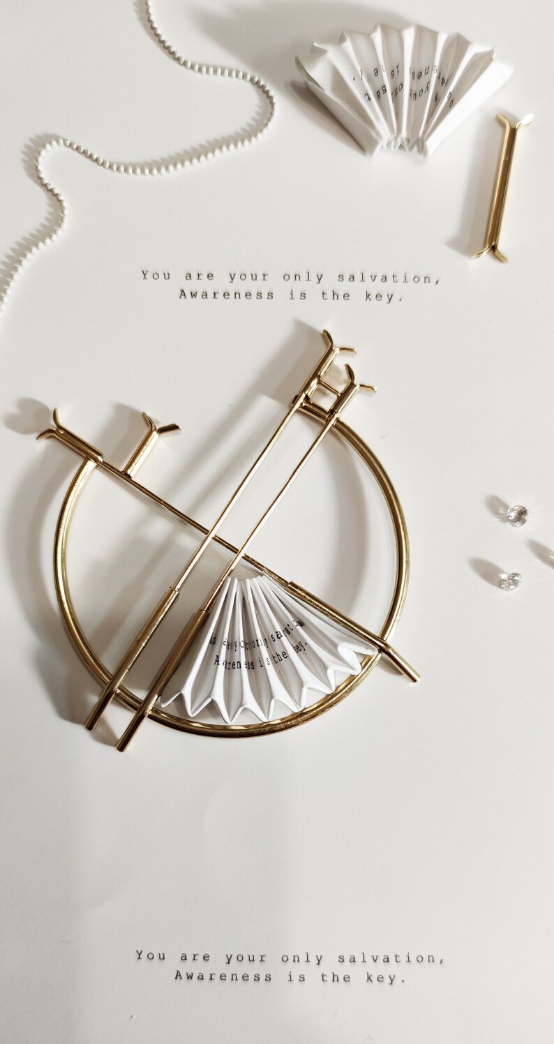 Puede incluir: Un soporte de metal dorado con una parte superior redonda y dos brazos largos. Un abanico de papel blanco se coloca en el soporte. El abanico tiene el texto "You are your only salvation, Awareness is the key." escrito en &eacute;l.
