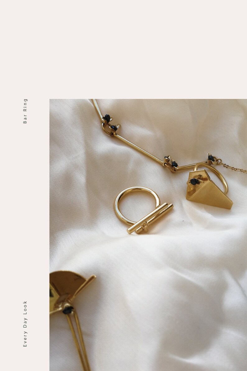 May include: Three gold rings with black stones. The rings are on a white fabric background. The rings are a bar ring, a triangle ring, and a half circle ring. The text "Every Day Look" and "Bar Ring" are on the left and right sides of the image.