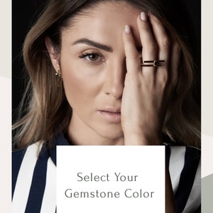May include: A close-up of a woman's hand wearing a gold ring with a black gemstone. The text "Select Your Gemstone Color" and "Black / Clear" is displayed below the image.