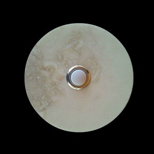 Könnte beinhalten: Ein runder, cremefarbener Türklingelknopf mit silbernem Rand und einem weißen Knopf in der Mitte.