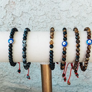May include: Five beaded bracelets with an evil eye charm. The bracelets are displayed on a white stand. The bracelets are made of black onyx, tiger's eye, and gray stones.