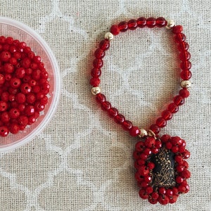 May include: A red beaded necklace with a red fabric pendant. The pendant has a gold design and is shaped like a rectangle.