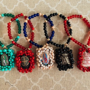 May include: Five colorful beaded bracelets with religious charms. Each charm is a different color and features a different religious image. The charms are surrounded by beads in the same color as the charm.