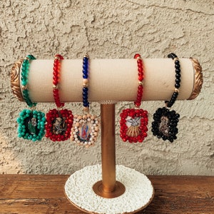 May include: Five beaded bracelets with religious charms. Each bracelet is a different color: green, red, blue, red, and black. The charms are square and feature embroidered images of religious figures.