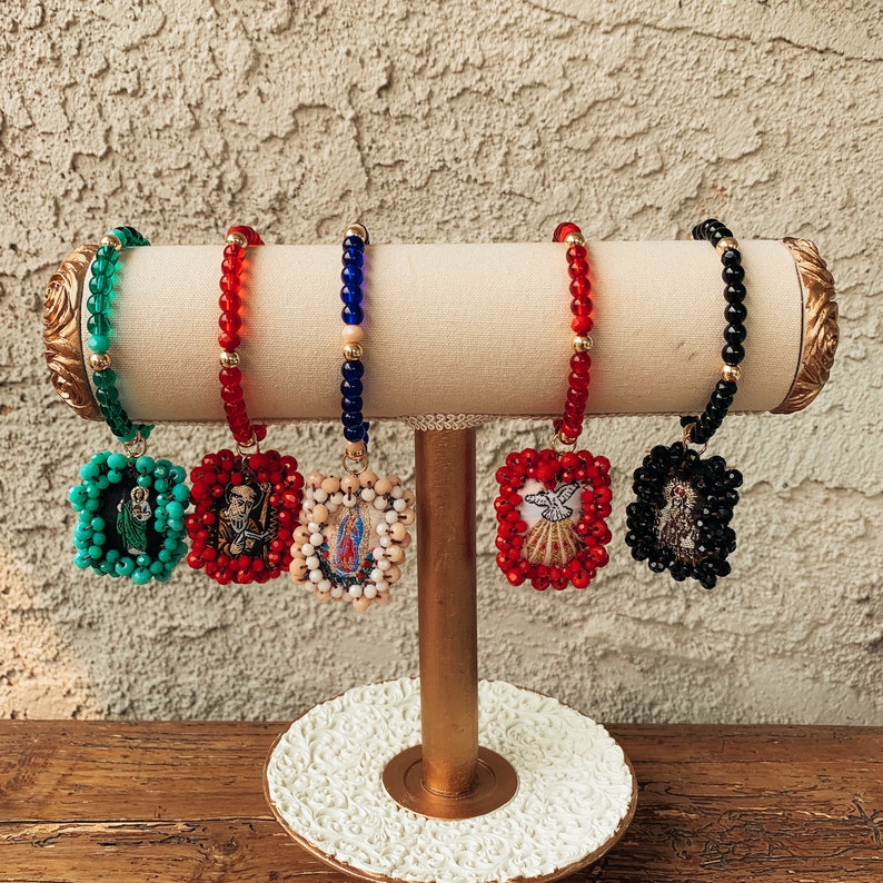May include: Five beaded bracelets with religious charms. Each bracelet is a different color: green, red, blue, red, and black. The charms are square and feature embroidered images of religious figures.