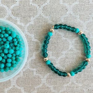 May include: A teal blue bracelet made of glass beads with silver accents. The bracelet is on a light brown fabric background.