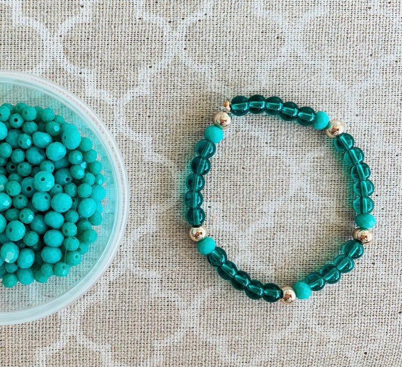 May include: A teal blue bracelet made of glass beads with silver accents. The bracelet is on a light brown fabric background.