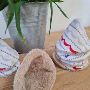 May include: A stack of white, patterned reusable facial rounds with a wave and dot design in red, blue, and gray. One round is shown with a tan terry cloth side. A galvanized metal pot with green plants is in the background.