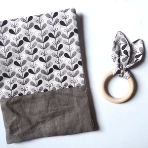 May include: A baby blanket and a wooden teether. The blanket has a white background with a black and gray leaf pattern and a dark gray bottom. The teether has a wooden ring with a fabric bunny ear design in matching colors.