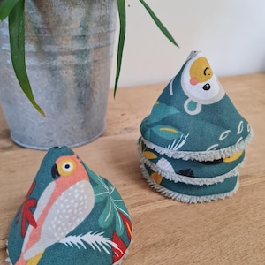 May include: Two teal fabric pads with parrot and tropical leaf designs. One pad is separate, and the other pads are stacked. The pads have a white, fluffy trim. A potted plant in a metal bucket is in the background.