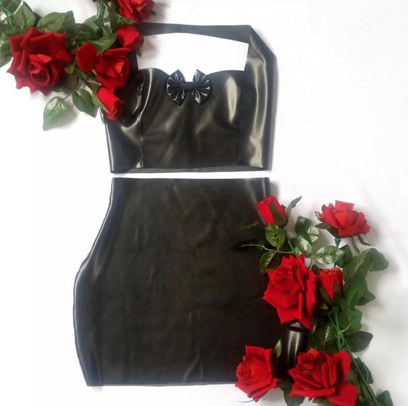 May include: Black latex two-piece outfit featuring a halter top with a bow detail and a matching mini skirt. The outfit is displayed on a white background and is accented with red roses.