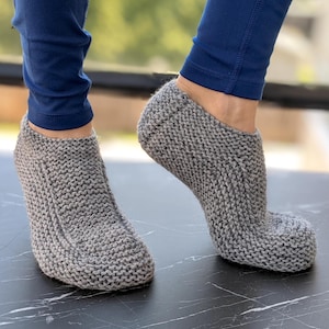 Pode incluir: Um par de meias de pantufa cinzentas de malha. As meias são feitas de um fio espesso e texturizado e têm uma biqueira arredondada e um punho canelado. As meias são mostradas numa superfície preta.