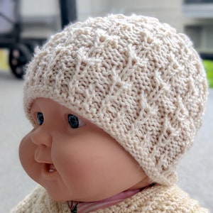 May include: A knitted baby hat in a cream color with a cable knit pattern. The hat is on a baby doll's head.
