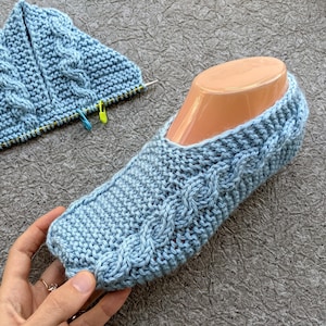 Puede incluir: Un par de pantuflas de punto azul con un patrón de trenza. Las pantuflas están siendo sostenidas por una mano.