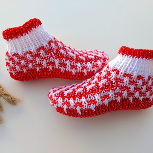 May include: A pair of knitted red and white patterned booties. The booties have a red cuff and a repeating pattern of red and white stitches. The booties are designed to keep feet warm and cosy.