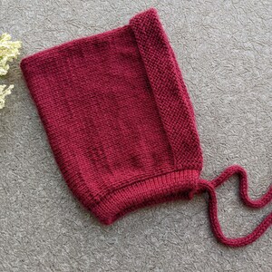 Może przedstawiać: Dzianinowy bordowy czepek dla dziecka ze sznurkami. Czepek wykonany jest z miękkiej, teksturowanej przędzy i posiada składany brzeg. Sznurki są długie i można je zawiązać pod brodą. Czepek jest w jednolitym kolorze.