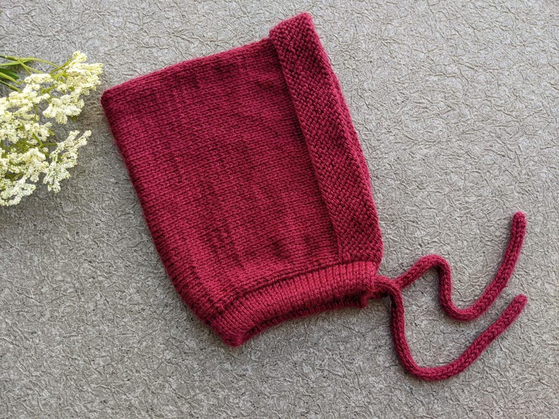 Może przedstawiać: Dzianinowy bordowy czepek dla dziecka ze sznurkami. Czepek wykonany jest z miękkiej, teksturowanej przędzy i posiada składany brzeg. Sznurki są długie i można je zawiązać pod brodą. Czepek jest w jednolitym kolorze.