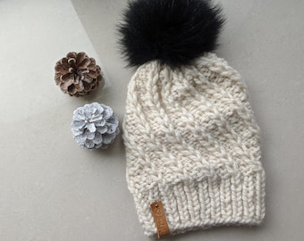 May include: A white knitted beanie with a black faux fur pom pom. The beanie has a cable knit pattern and a brown leather tag that says "The Life".